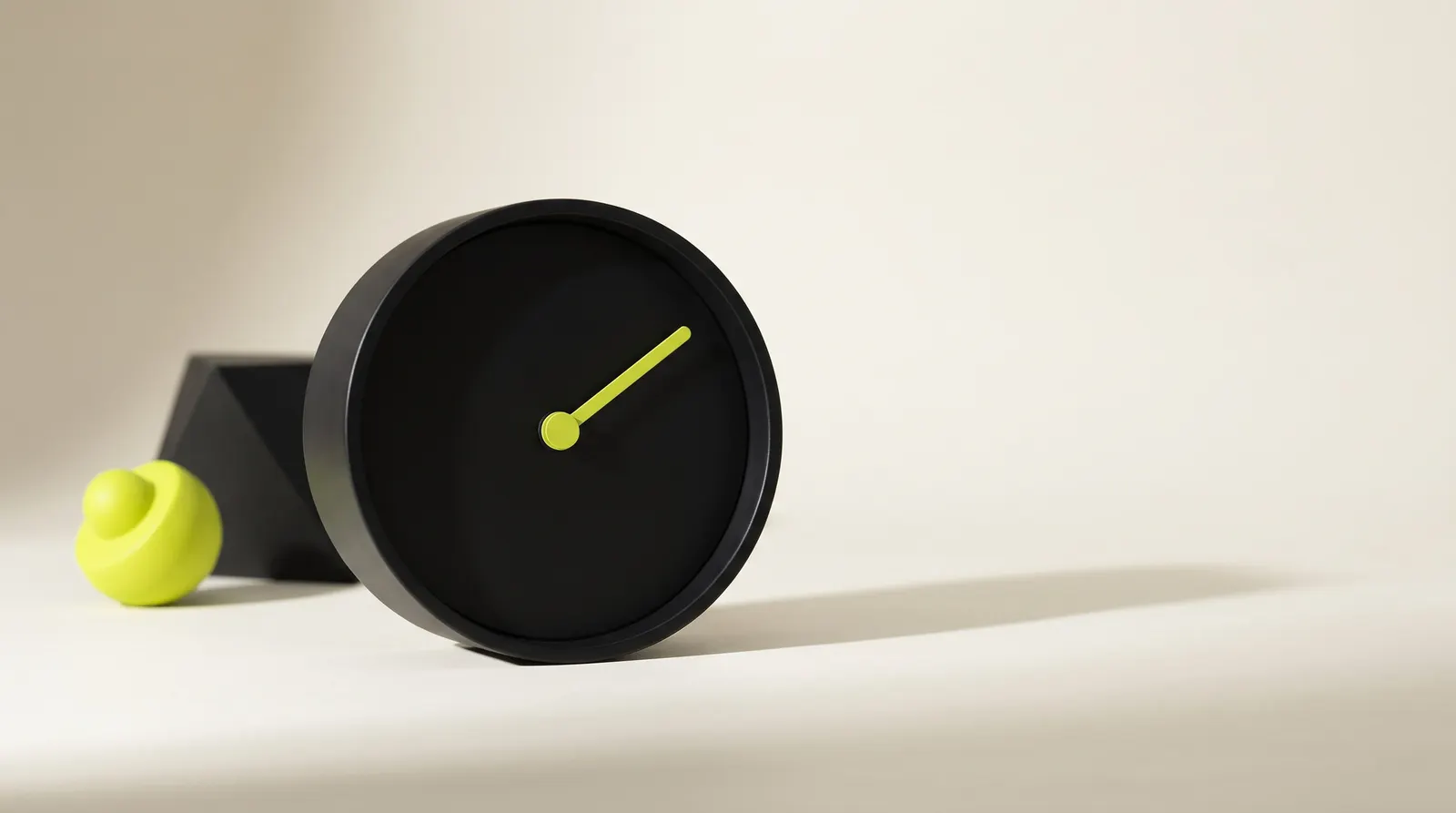 Reloj de escritorio negro con la esfera lisa y una sola aguja verde lima, junto a un bloque negro.
