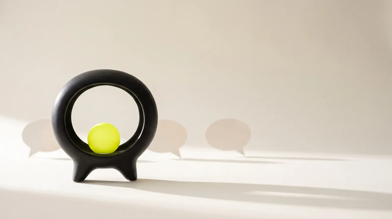 Black ring-shaped sculpture on a pale surface holding a lime sphere, with paper speech bubbles standing behind it.