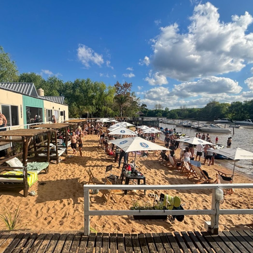 Busy riverside beach in the Delta with white umbrellas, tables and deck chairs, and boats moored along the shore